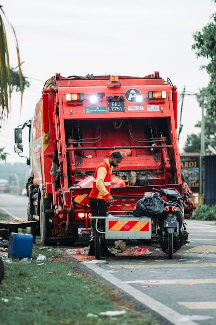 A worker wearing a high-visibility yellow and orange vest is attending to a large, red waste collection vehicle parked on the side of a street. The vehicle, used for rubbish removal, features a prominent rear compactor with visible hydraulic components and safety markings. The worker is leaning over the open rear chamber, likely managing waste or preparing for collection. Next to the vehicle, there is a small black motorcycle with a black plastic bag placed on the seat, indicating it may be part of a waste collection process or nearby personal transport. The street environment includes a paved sidewalk, a patch of grass, and a few scattered debris such as plastic bottles and paper beneath the vehicle and near the curb. In the background, there are trees, utility poles, and buildings, suggesting an urban or suburban setting. The scene is lit by natural daylight, with soft, diffused lighting. The image exemplifies on-site waste management activities, aligning with private rubbish collection services, and highlights the practical aspects of waste clearance operations in Surrey.