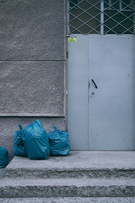 A row of four blue plastic rubbish bags are placed on a concrete step in front of a metal door with a black handle, situated against a grey textured wall on the left side of the image. The wall features a vertical edge dividing the surface, and the door is part of a larger metal structure with a grid-like security grille above it. There is a small yellow and green warning sticker affixed near the top of the door frame. The ground appears to be pavement, and the overall environment suggests an urban or industrial setting. The scene relates to waste collection and disposal, as the bags are likely prepared for rubbish removal services, with Waste Clearance Surrey possibly providing such an independent solution for waste management and alternative disposal methods in the area. Natural lighting highlights the textures of the concrete, wall, and bags, emphasizing their materials and placement within this typical rubbish storage area.