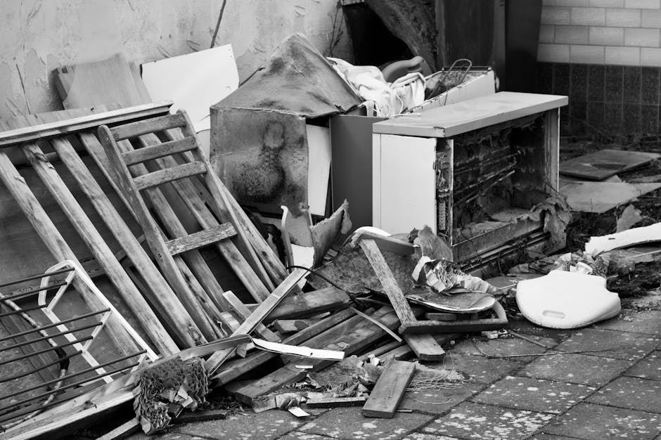 A cluttered outdoor scene showing a disorganized pile of discarded household items and waste, including a wooden ladder with multiple rungs leaning against a wall, and various debris scattered across a paving surface. The debris consists of broken wooden planks with rough, splintered edges, a damaged white refrigerator door with visible rust and dirt, and a piece of white plastic piping. There is also a white, rounded plastic object, possibly a toilet seat, lying on the ground. Behind the pile, a brick and concrete wall forms part of the environment, while the area appears to be an alleyway or back yard used temporarily for waste accumulation. The overall scene is in black and white, emphasizing the textures of the materials, the weathered surfaces, and the disorder associated with non-organic rubbish awaiting professional clearance from a service such as Waste Clearance Surrey, highlighting the need for proper disposal or removal services for bulky waste.