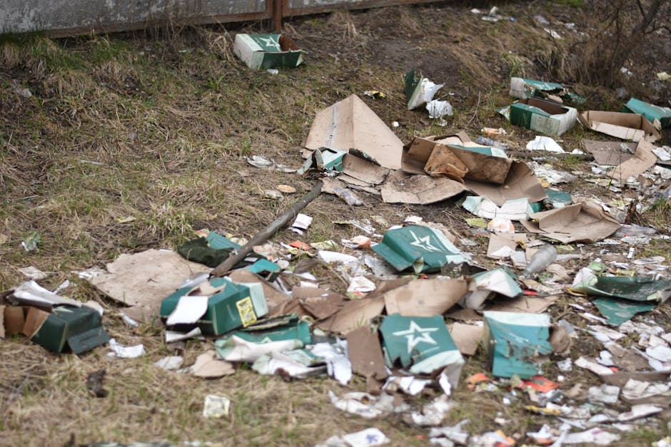 A cluttered outdoor scene showing a disorganized pile of discarded household items and waste, including a wooden ladder with multiple rungs leaning against a wall, and various debris scattered across a paving surface. The debris consists of broken wooden planks with rough, splintered edges, a damaged white refrigerator door with visible rust and dirt, and a piece of white plastic piping. There is also a white, rounded plastic object, possibly a toilet seat, lying on the ground. Behind the pile, a brick and concrete wall forms part of the environment, while the area appears to be an alleyway or back yard used temporarily for waste accumulation. The overall scene is in black and white, emphasizing the textures of the materials, the weathered surfaces, and the disorder associated with non-organic rubbish awaiting professional clearance from a service such as Waste Clearance Surrey, highlighting the need for proper disposal or removal services for bulky waste.