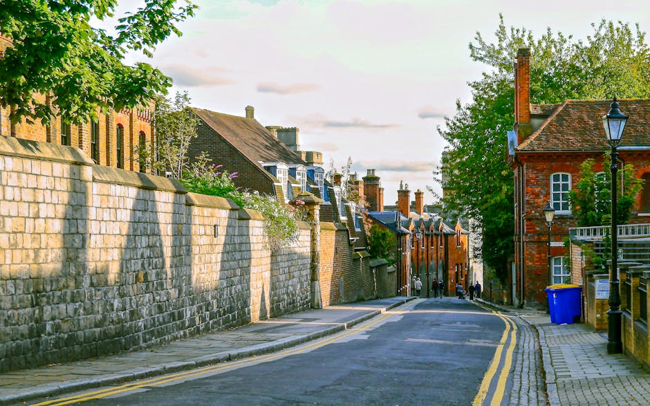 A quiet street scene featuring a narrow asphalt road with double yellow lines running parallel to a stone pavement on the right side. The left side of the street is bordered by a high, weathered stone wall with a rough texture, casting shadows on the pavement. Behind the wall, several brick buildings with varying architectural styles and slate or tiled roofs are visible, some with multiple chimneys and white window frames. To the right, there is a red brick building with white-framed windows, a small balcony, and a traditional black lamppost positioned near the sidewalk. A blue recycling bin stands against the building's wall, and behind it are mature trees with lush green foliage, indicating a spring or early summer season. In the distance, a small group of pedestrians can be seen walking along the sidewalk. The overall scene is illuminated by soft, natural daylight, showcasing a typical residential street that may be used for private waste collection or alternative rubbish removal services, as suggested by the company's presence in the local area of Surrey, UK.