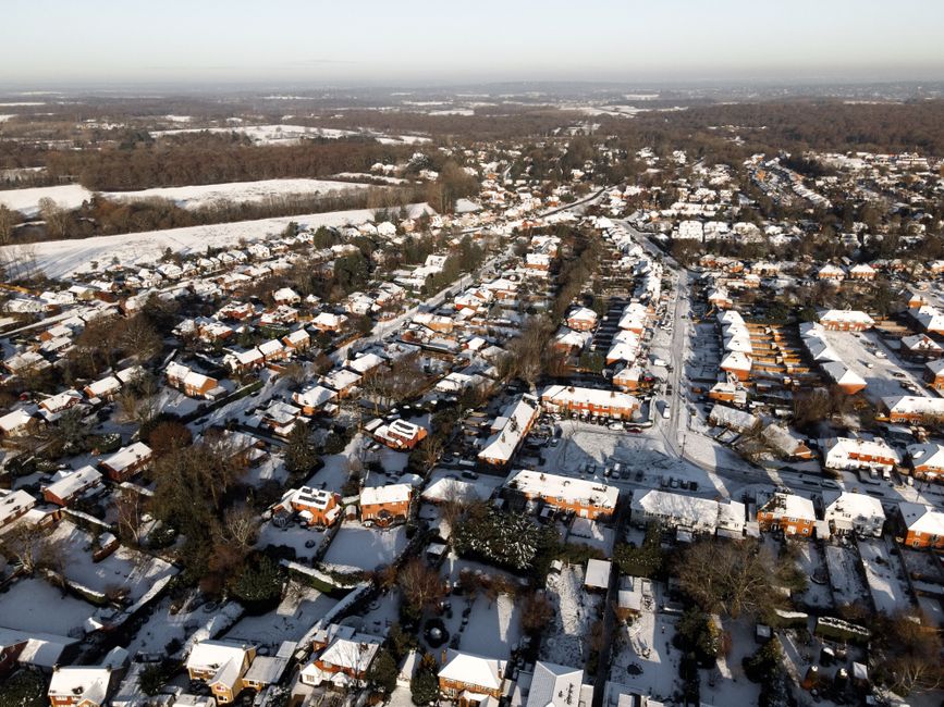 An aerial view of a residential suburban area covered in snow, showing rows of terraced and semi-detached houses with reddish-brown brick exteriors and white snow on their pitched roofs. Streets are lined with parked cars, some partially obscured by snow, and the roads themselves have visible snow coverage. Scattered trees, some bare and some with snow-dusted foliage, are interspersed throughout the neighborhood. In the background, expansive open fields and woodland areas are also blanketed in snow, extending towards the horizon under a pale, overcast sky. The scene is captured during daylight, with diffuse natural light creating soft shadows. This type of environment may be typical for private waste clearance or rubbish removal services, reflecting the need for local, independent collection options offered by waste management providers such as Waste Clearance Surrey, particularly in areas where snow impact may delay municipal rubbish collection services.